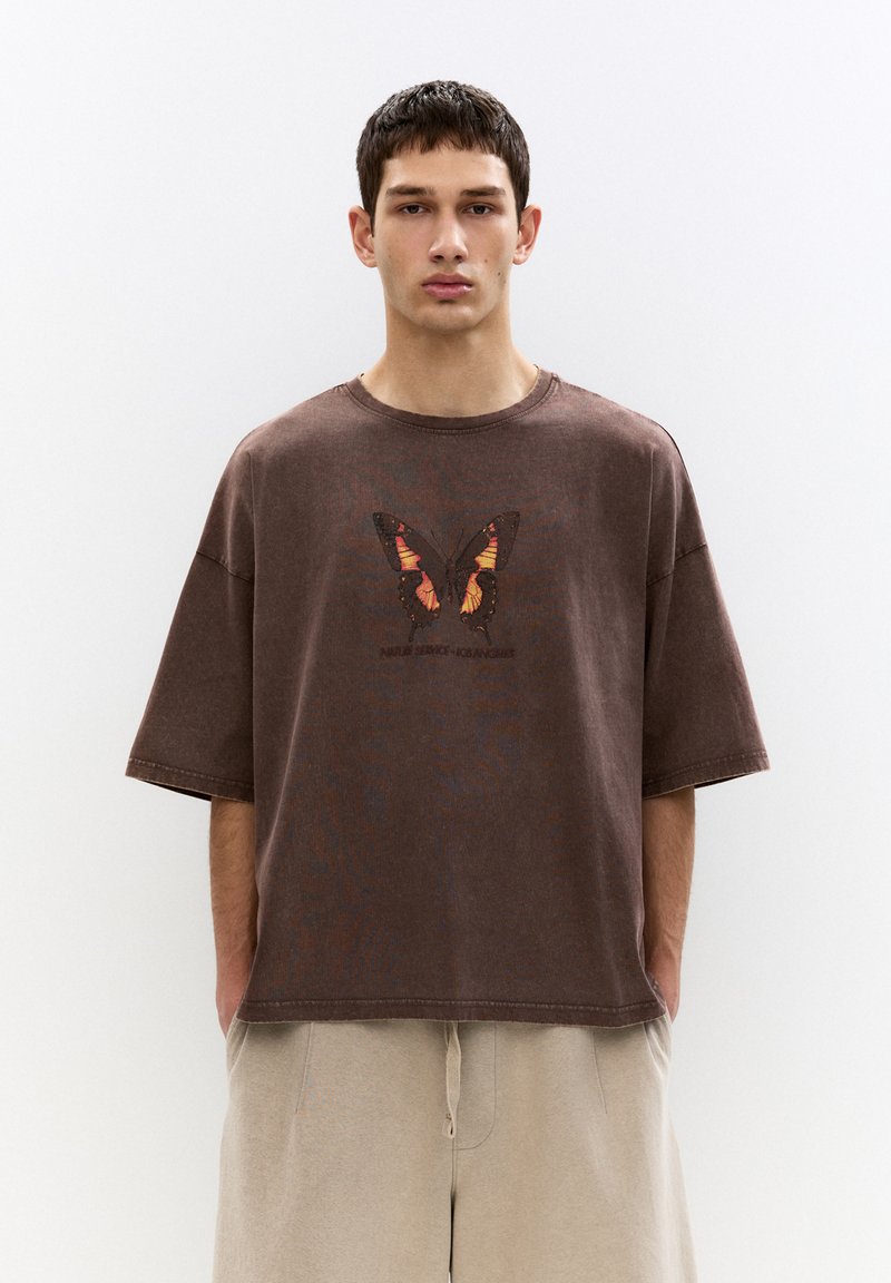 T-shirt oversize marrone con grafica di una farfalla in arancione e nero, con scollatura rotonda e dettagli delle cuciture in vista.
