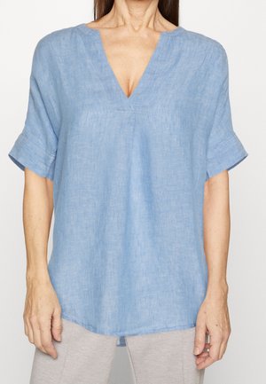 Femme portant un haut ample en lin bleu clair à manches courtes avec un col en V et des manches retroussées, associé à un pantalon gris clair.