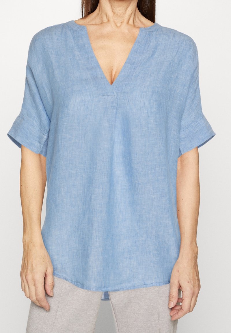 Femme portant un haut ample en lin bleu clair à manches courtes avec un col en V et des manches retroussées, associé à un pantalon gris clair.