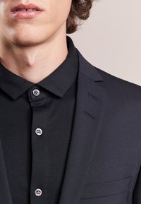 Black blazer with a standard collar, single button closure, and two visible pockets. Smooth fabric texture and contrasting buttons.