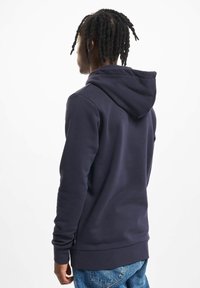 Mænd med fletninger iført en mørk navy hoodie og blå jeans, som står med ryggen til en simpel hvid baggrund.
