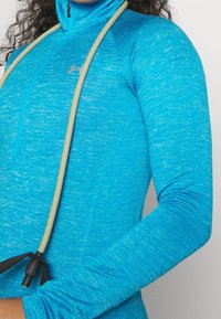 Modrý atletický top s dlouhým rukávem a čtvrtovým zipem, vyrobený z texturované látky. Má světle zbarvený úvazek připevněný u krku.