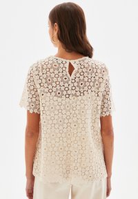 Haut en dentelle crème avec motifs floraux, manches courtes et ouverture en forme de goutte dans le dos. Design en crochet texturé sur toute la surface.