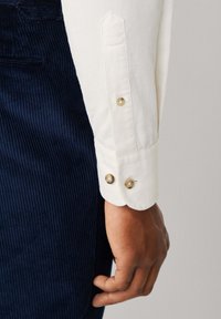 Witte langemouwen overhemdmanchet met twee gouden knopen, gecombineerd met een gestructureerde marineblauwe corduroy stof.