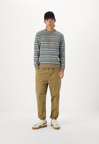 El suéter de punto presenta un patrón geométrico azul, complementado por pantalones beiges. Una gorra negra y zapatillas blancas con suelas marrones completan el look.