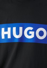 Schwarzes Baumwoll-T-Shirt mit einem auffälligen blauen rechteckigen Grafikdesign, auf dem in großen weißen Buchstaben der Text "HUGO" auf der Vorderseite steht.
