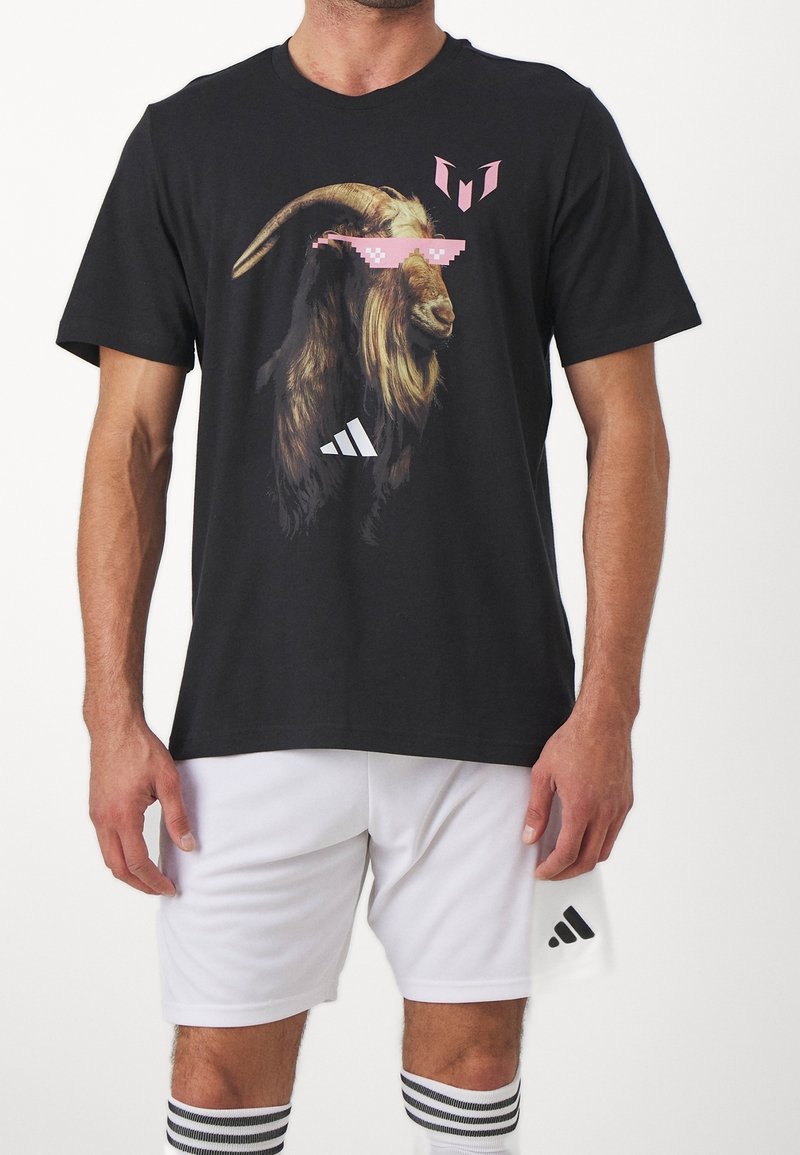 Mustat puuvillapaita, jossa on vuohigrafiikka, jossa on vaaleanpunaiset pikselöidyt aurinkolasit ja valkoiset Adidas-logot etupuolella, yhdistetty valkoisiin shortseihin.