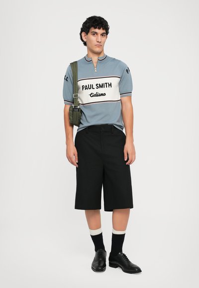 Joven con una camiseta de ciclismo azul de Paul Smith, pantalones cortos largos negros, calcetines negros con bandas blancas, zapatos de vestir negros y una bolsa cruzada verde oliva.