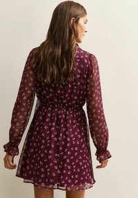 Vestido floral burdeos con mangas largas transparentes, cintura elástica y puños con volantes; presenta pequeñas flores rosas sobre un tejido texturizado.