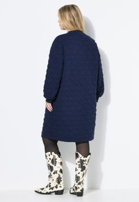 Marineblaues Steppkleid mit langen Ärmeln und rundem Ausschnitt, kombiniert mit kniehohen weißen Stiefeln mit einem schwarz-braunen Muster.