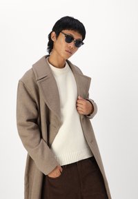 Abrigo de lana marrón con grandes solapas, suéter de canalé crema y pantalones de pana marrones. Modelo usando gafas de sol negras, posando con una mano en el abrigo.