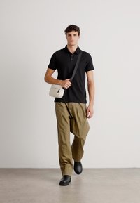 Čierna polokošeľa, voľné olivovo zelené nohavice a čierne topánky. Model má na sebe malú smotanovú crossbody tašku s popruhom. Hladké textúry.