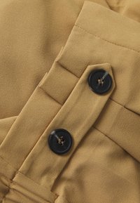 Tessuto color sabbia con una texture liscia, caratterizzato da due bottoni neri e dettagli piegati, che mostrano le cuciture su una sezione del capo.