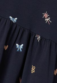 Tela de algodón azul marino con patrones bordados de lazos, corazones, estrellas y flores en azul claro, rosa y amarillo. Diseño de dobladillo acampanado.