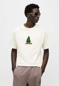 Εκρού κοντομάνικη μπλούζα με πολύχρωμο γραφικό δέντρο και κείμενο «Converse»· συνδυάζεται με καρό σο ρτς σε πορτοκαλί και μπλε αποχρώσεις.