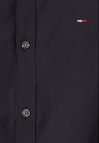 Chemise noire à boutons avec un petit logo rouge, blanc et bleu, texture lisse et deux boutons noirs le long de la fermeture.