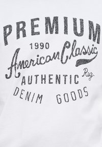 Λευκό βαμβακερό t-shirt με μαύρα, υφασμένα γραφικά γράμματα που περιλαμβάνουν τις λέξεις «PREMIUM», «American Classic» και «AUTHENTIC DENIM GOODS» με ένα vintage σχέδιο.