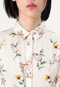 Leichtes, blumig gemustertes Hemd mit weißem Hintergrund. Verfügt über einen klassischen Kragen, Knopfverschluss und farbenfrohe botanische Akzente.