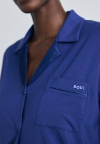 Chemise bleu à boutons fabriquée en tissu lisse avec un col, présentant une poche avant avec le mot "BOSS" brodé en blanc.