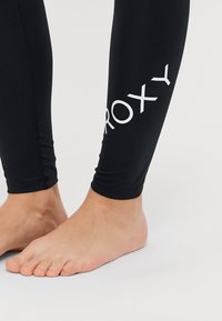 Leggings neri realizzati in tessuto elasticizzato, con un logo "ROXY" bianco sul lato e un design aderente. Piede mostrato con le dita a piedi nudi.