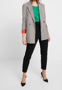 Gray houndstooth blazer with orange cuffs, paired with a green blouse and black fitted trousers. Model wearing black heels.