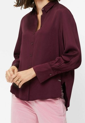 Woman wearing a long-sleeve dark purple button-up shirt with a collar and light pink corduroy pants, hands lightly clasped in front.