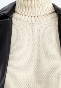 Beige gestrickter Rollkragenpullover mit gerippter Textur, kombiniert mit einer schwarzen Lederjacke, die auf der linken Seite des Bildes sichtbar ist.