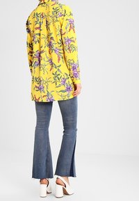 Camicia lunga gialla a fantasia floreale con colletto, abbinata a jeans svasati blu scuro con spacchi laterali e scarpe bianche con tacco a blocco.