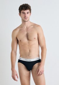 Fekete pamut bugyi fehér Calvin Klein logós derékpánttal. A dizájn sima textúrával és illeszkedő formával rendelkezik. Minimalista stílus.