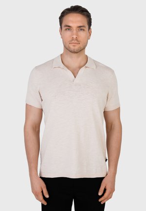 Mann som har på seg en lys beige kortermet polotrøye med krage, stående mot en ensfarget hvit bakgrunn.