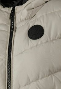 Ljusbeige, kviltad pufferjacka med svart dragkedja, prydd med en rund svart logotyp med text och geometrisk design. Slät yta.