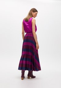 Top senza maniche in satin magenta abbinato a una gonna maxi plissettata con righe orizzontali viola e blu navy. Stivali borgogna fino al ginocchio.