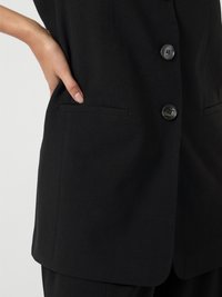 Colete preto sob medida com um design estruturado, apresentando três botões, uma textura suave e bolsos laterais.