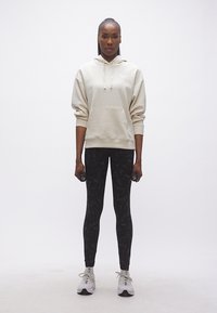 Γυναίκα με μπεζ φούτερ με κουκούλα και μαύρα κολάν κρατώντας βαράκια 1 κιλού, όρθια μπροστά σε απλό λευκό φόντο.