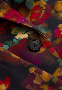 Látka s květinovým vzorem v živých barvách včetně červené, žluté a zelené, s černým pozadím; doplněno jediným černým knoflíkem.