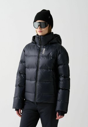 Zwarte pufferjack met een ritssluiting aan de voorkant, capuchon en een glanzende, gewatteerde textuur. Gecombineerd met een zwarte gebreide beanie en oversized zonnebril.