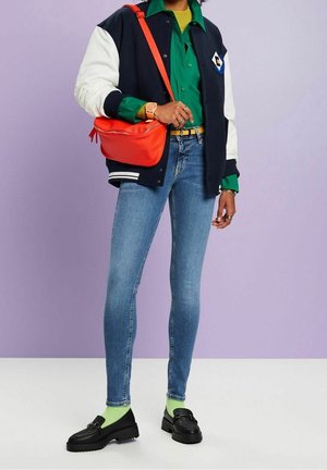 Giacca varsity blu navy e bianca, camicia verde con bottoni, jeans skinny blu, mocassini neri, borsa rossa brillante e calzini verde neon.