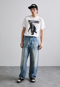 Fehér póló fekete gorilla grafikával és "MOSCHINO" felirattal, laza kék farmernadrággal és fekete slip-on cipőkkel párosítva. Fekete sapka is included.