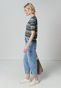 Femme blonde de profil portant un haut à manches courtes à motifs, un jean bleu clair, des baskets blanches, tenant un sac à bandoulière marron sur un fond uni.