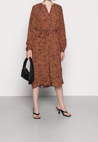 Abito lungo a maniche lunghe con stampa leopardata marrone, scollatura a V, vita arricciata e tessuto fluido. Abbinato a una borsa a tracolla nera intrecciata e sandali neri con tacco.