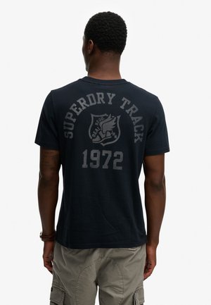 Mann mit kurzen lockigen Haaren, der ein dunkelblaues Superdry Track 1972 T-Shirt und hellfarbene Cargo-Shorts trägt, von hinten gesehen.