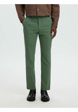 Mężczyzna w dopasowanych zielonych spodniach, brązowej, długorękawowej koszuli włożonej do środka oraz czarnych skórzanych mokasynach, stojący z rękami częściowo w kieszeniach.