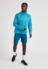Maglia sportiva azzurro chiaro con zip e maniche lunghe, logo sul petto, abbinata a pantaloni corti color teal e scarpe da ginnastica nere con dettagli bianchi.