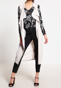 Long gilet sans manches en beige clair avec un col drapé. En dessous, un top noir à motifs floraux et un pantalon noir ajusté. Talons ouverts noirs.