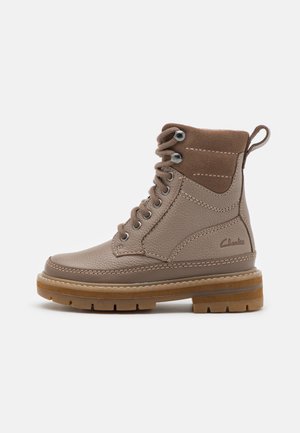 Bottine en cuir marron à lacets avec col rembourré, semelle épaisse en caoutchouc et languette au talon, affichant le logo Clarks sur le côté.