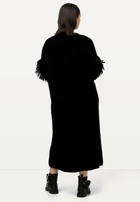 Cardigan lungo nero in tessuto testurizzato, con frange sui polsini. Indossato sopra stivali neri alla caviglia.