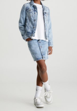 Persoon draagt een lichtblauwe spijkerjasje en rok, wit T-shirt, witte Calvin Klein sokken en witte sneakers, staat met één been gekruist.