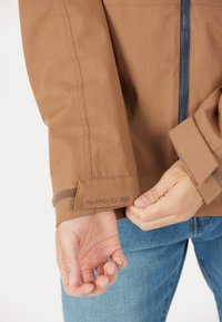 Brun vattentät jacka med dragkedja, med en texturerad mudd och broderad "WPRO 10000" detalj. Bärs med blå jeans.