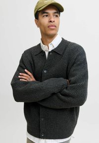 Mørkegrå ribstrikket cardigan med kravehals, knapper og løs pasform. Båret over en hvid skjorte, hvilket fremhæver en tekstureret overflade.