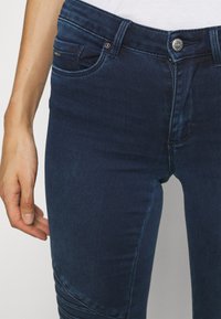 Vaqueros de mezclilla de un azul oscuro con un diseño ajustado, cinco bolsillos y cremallera frontal. Presenta un botón de metal y detalles de costura sutiles.